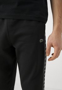 Sorte joggingbukser lavet af glat stof, med et lille grønt krokodillelogo og en sideliste med gentaget tekst i hvid.