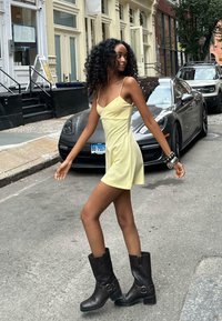 Yellow mini dress with spaghetti straps, paired with black leather ankle boots. Background features a city street with parked cars.