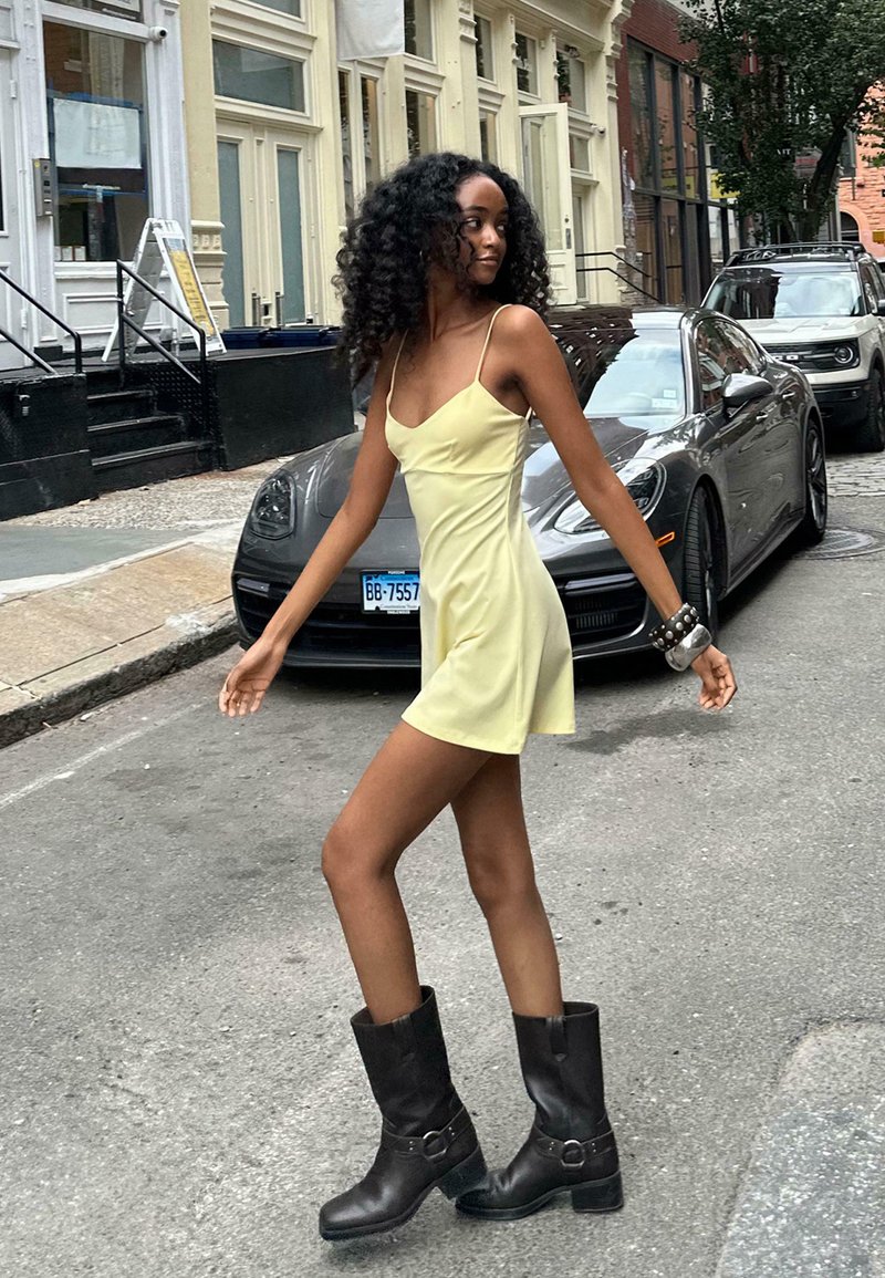 Yellow mini dress with spaghetti straps, paired with black leather ankle boots. Background features a city street with parked cars.