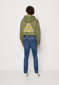 Olivgrön hoodie med en grafisk design på baksidan, i kombination med blå slim-fit jeans och vita sneakers med grå detaljer.