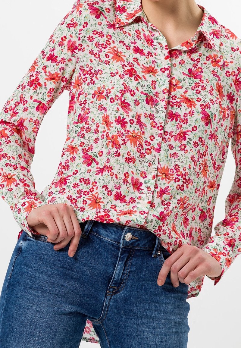 Chemise à fleurs à boutons, présentant un fond blanc, des fleurs roses et rouges, des manches longues et un col classique. Associée à un jean en denim bleu.