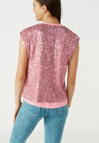 Débardeur sans manches en tissu rose orné de sequins, avec un col rond et un ourlet rose contrasté. Associé à un jean bleu clair.