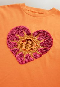 Chemise orange avec un motif de soleil en forme de cœur composé de sequins réversibles roses et dorés sur la poitrine.