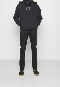 Marinblå hoodie med snoddar och en logotyp på framsidan, kombinerad med svarta byxor och svarta sneakers med gummisålar och vita accenter.