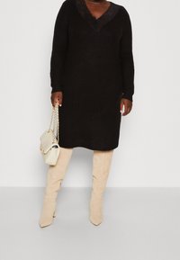 Černé pletené šaty s dlouhými rukávy a výstřihem do V zdobeným krajkou. Nosit je béžové vysoké boty nad kolena a držet krémovou kabelku.