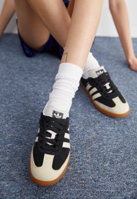 Black and beige sneakers with a suede and leather upper, featuring three white stripes, black laces, and gum rubber sole.