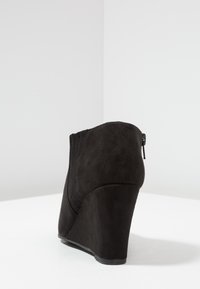 Black wedge ankle boot made of suede, featuring a smooth texture, zipper closure, and rounded toe design.