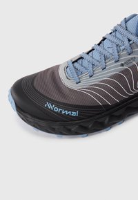 Nahaufnahme eines schwarz-blauen Sportschuhs mit strukturiertem Sohlen, blauen Schnürsenkeln, weißen Wellenmustern und Gore-Tex Invisible Fit-Label an der Seite.