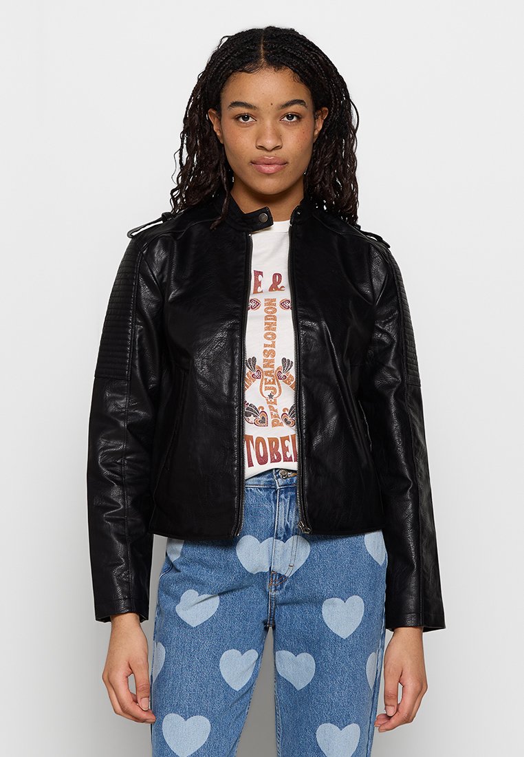 Mujer con el cabello trenzado que lleva una chaqueta de cuero negra, una camiseta gráfica blanca y unos jeans azules con patrones de corazones blancos, sobre un fondo blanco.