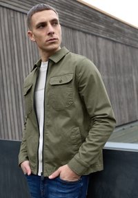 Kurtka na zamek w kolorze oliwkowej zieleni z dwoma kieszeniami na piersi, zestawiona z beżowym swetrem i niebieskimi jeansami; gładki materiał i usztywniony krój.