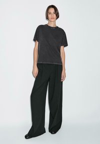 T-shirt nera oversize in cotone con scollatura rotonda, abbinata a pantaloni neri ampi e plissettati e scarpe nere su uno sfondo neutro.