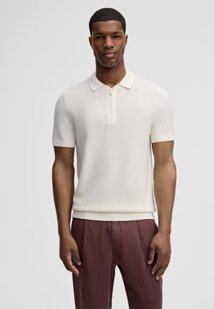 Mann trägt weißes Strick-Poloshirt mit kurzen Ärmeln und Reißverschluss sowie weinrote Hose, steht vor schlichtem grauem Hintergrund.