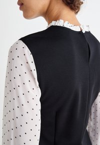 Musta koru, jossa on teksturoitu pinta, yhdistetty valkoisiin läpikuultaviin hihoihin, joissa on mustia pilkkuja, sekä röyhelökaulukseen. Napitus takana.