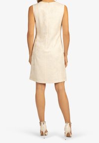 Bezramenné krémové šaty s texturovaným květinovým vzorem, s přiléhavým střihem a zipem na zadní straně. Noseny s béžovými jehlovými botami s kotníkovými pásky.