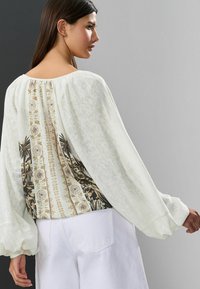 Blouse blanche avec des manches amples, présentant un dos à motifs beige, noir et doré. Tissu texturé avec une coupe décontractée.