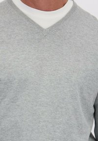 Man in een lichtgrijze V-hals sweater over een witte ronde hals T-shirt, waarbij alleen de nek en het bovenste deel van de borst zichtbaar zijn.