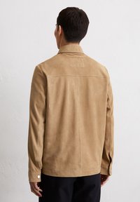 Tan suède jas met een kraag, lange mouwen en drukknopen. Glad textuur, minimalistisch ontwerp en subtiele stikseldetails.