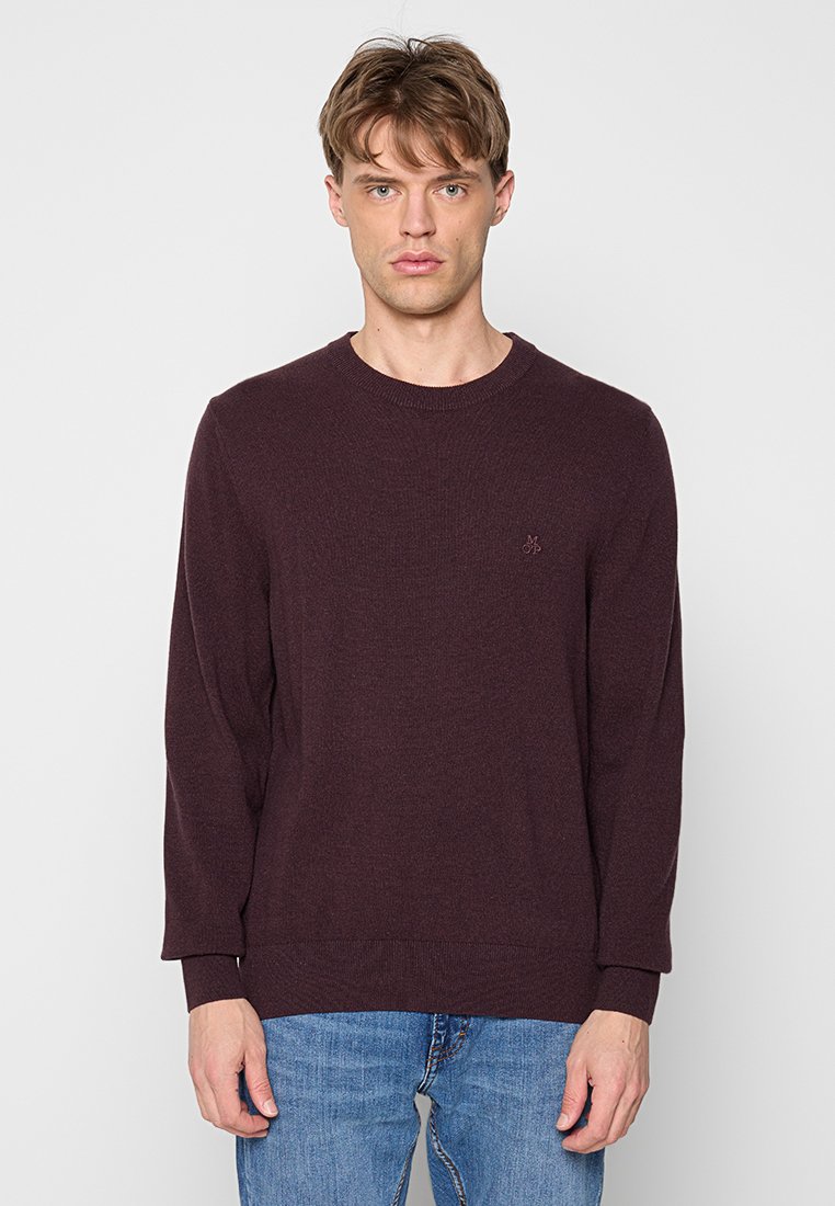 Junger Mann in einem dunkelvioletten Rundhalsausschnittpullover und blauen Jeans, der vor einem schlichten hellgrauen Hintergrund steht.