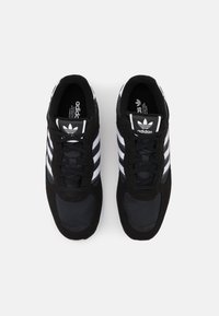 Černé sportovní boty s bílým designem se třemi pruhy, vyrobené z jemné kůže a síťoviny, kulatá špička a černé šněrování. Logo na jazyku.