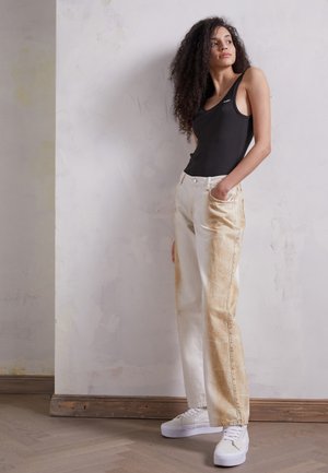 Jeans de denim desgastados brancos e bejes com um corte descontraído, combinados com uma camisola preta canelada e ténis brancos. Fundo em madeira e gesso.