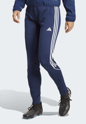 Person wearing navy blue Adidas track pants with white stripes and black soccer cleats, mid-step on a light background.