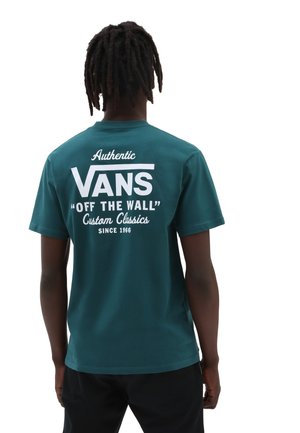Person mit dunkler Hautfarbe und Dreadlocks, von hinten gesehen, trägt ein türkises Vans-T-Shirt mit weißem Schriftzug "Authentic VANS Off The Wall Custom Classics Since 1966".