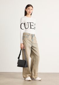Chemise blanche à manches longues avec texte noir, jeans larges en denim clair, sac bandoulière noir et baskets blanches avec des accents clairs.