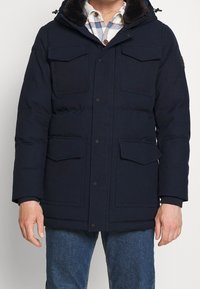 Hombre vestido con una chaqueta de invierno acolchada azul marino con cuatro bolsillos frontales de solapa sobre una camisa de cuadros blanca y azul y jeans azul.