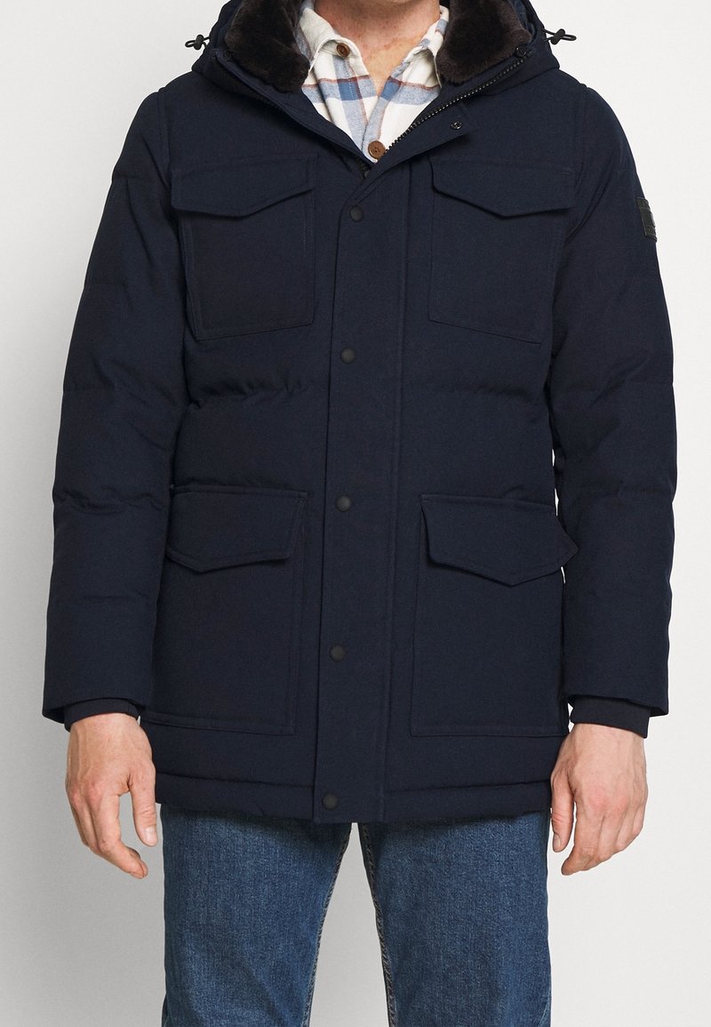 Hombre vestido con una chaqueta de invierno acolchada azul marino con cuatro bolsillos frontales de solapa sobre una camisa de cuadros blanca y azul y jeans azul.