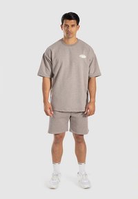Graues Baumwoll-T-Shirt mit kurzen Ärmeln und kleinem Logo, kombiniert mit passenden Shorts. Das Modell trägt weiße Turnschuhe und gestreifte Socken.