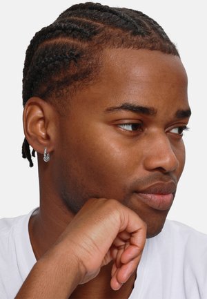 Nahaufnahme einer Person mit geflochtenem Haar und einem silbernen Creolenohrring, die mit dem Kinn auf der Hand ruht und ein weißes T-Shirt trägt, vor einem schlichten Hintergrund.