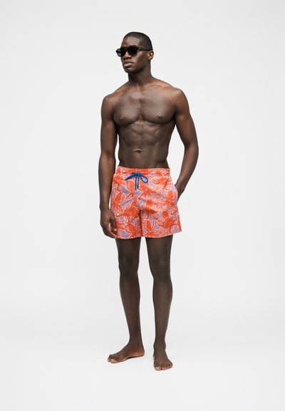 Άνδρας με μαύρα γυαλιά ηλίου και πορτοκαλί μαγιό με μπλε και λευκά σχέδια παϊνλεϊ, στέκεται ξυπόλητος σε απλό λευκό φόντο.