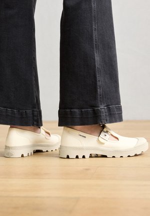 Beżowe płócienne buty z wycięciami i detalami w postaci paska, z teksturowanymi gumowymi podeszwami i widocznym logo z boku.