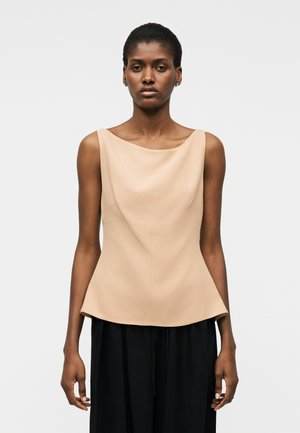 Femme portant un haut beige sans manches avec un col bateau et un pantalon noir ample, debout devant un fond clair uni.