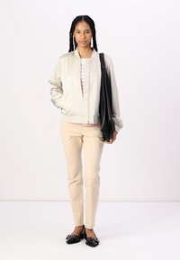 Giacca bomber grigia chiara con chiusura a zip, abbinata a una camicia a righe rosa, pantaloni beige e scarpe basse nere. Borsa nera appesa alla spalla.