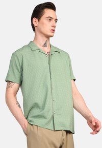 Camisa de botones de manga corta en verde claro con un patrón texturizado y cuello, con costuras blancas y botones grises.