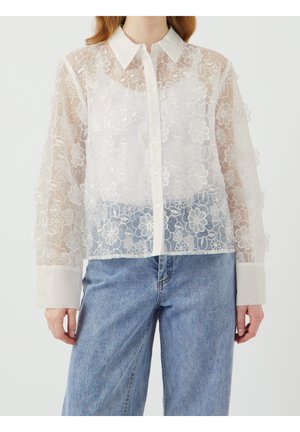 Chemise blanche en voile transparent avec broderie florale, manches longues, devant à boutons et col net. Associée à un jean en denim bleu.