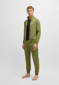Olijfgroen trainingspak met een jack met ritssluiting en bijpassende broek. Het jack heeft een hoge kraag en zijzakken; de stof lijkt glad en flexibel.
