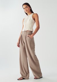 Crop top in tessuto beige chiaro con cerniera frontale, abbinato a pantaloni beige a vita alta e larghi e semplici sandali neri.
