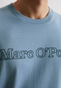 Blauwe katoenen t-shirt met een ronde hals, voorzien van een grote grijze decoratieve print van "Marc O'Polo" over de borst. Sterke textuur.
