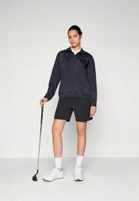 Tengerészkék szélálló dzseki fél cipzárral, elasztikus szegéllyel és hosszú ujjakkal; fekete rövidnadrággal és fehér sportcipővel párosítva, golfütőt tartva.