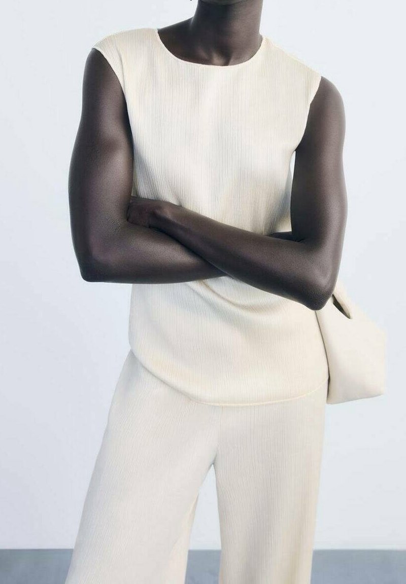 Personne à la peau foncée portant un haut crème sans manches et un pantalon assorti, les bras croisés, tenant un sac crème sur le côté gauche.