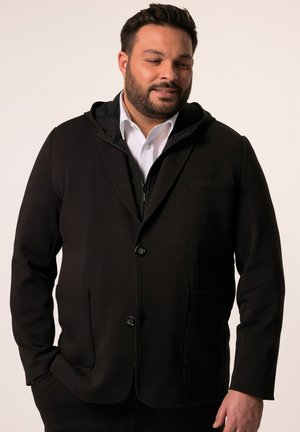 Schwarze Softshelljacke mit Kapuze, strukturiertem Design, zwei frontalen Taschen und Knopfverschluss. Über einem weißen Hemd mit Kragen getragen.