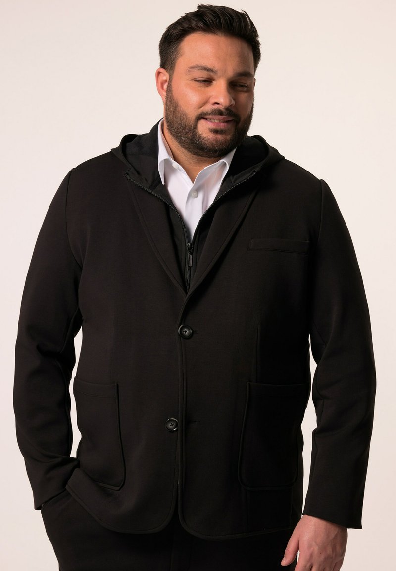 Giacca softshell nera con cappuccio, design strutturato, due tasche frontali e chiusura con bottoni. Indossata sopra una camicia bianca con colletto.