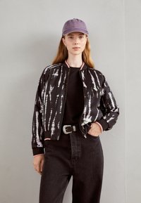 Veste bomber noire avec un motif tie-dye, accents roses, fermeture éclair, portée sur un haut noir et associée à un jean foncé taille haute et une casquette violette.