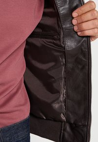 Dunkelbraune Lederjacke von innen mit einem glänzenden, glatten Futter und einer Reißverschlusstasche, mit gerippten Bündchen und Saum.