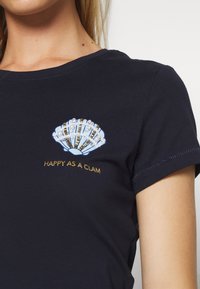 Námořnické bavlněné tričko s modrým vyšívaným motivem mušle se zlatými akcenty a textem "ŠŤASTNÝ JAKO MUŠLE" pod grafikou.