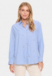 Camisa de botones a rayas azules y blancas con bordado, con herrajes en tono dorado, mangas largas y un ajuste relajado. Textura de tejido suave.