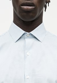 Κοντινό πλάνο ενός ατόμου που φορά ένα ανοιχτό μπλε καρό πουκάμισο με κουμπιά και μυτερό γιακά, δείχνοντας το λαιμό και το κάτω μέρος του προσώπου.