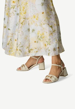 Sandales beiges à brides avec une texture tissée, talons carrés blocs, et des accents décoratifs en métal. Portées avec une robe d'été à motifs floraux.